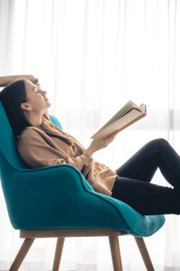 Çekici genç bir kadın, pencerenin yanındaki turkuaz koltukta oturmuş kitap okuyor, boşluğu kopyalıyor..