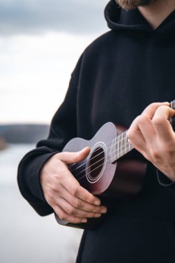 Genç bir adam ukulele gitarını doğada nehrin bulanık arka planında, rahatlama ve müzikal yaratıcılık kavramında çalar..