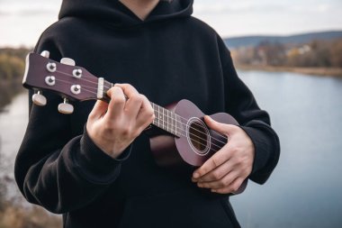 Genç bir adam ukulele gitarını doğada nehrin bulanık arka planında, rahatlama ve müzikal yaratıcılık kavramında çalar..