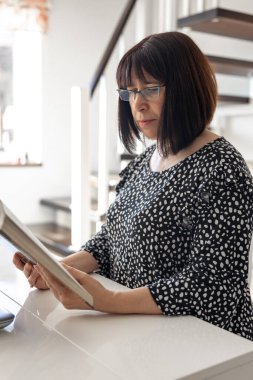 Gözlüklü orta yaşlı bir kadın evin içinde bir masada otururken kitap okur..