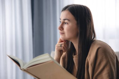 Genç bir kadın sabahları pencerenin yanındaki koltukta otururken kitap okur..