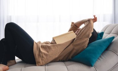 Genç bir kadın kanepeye uzanmış elinde bir kitapla fotokopi çekiyor..