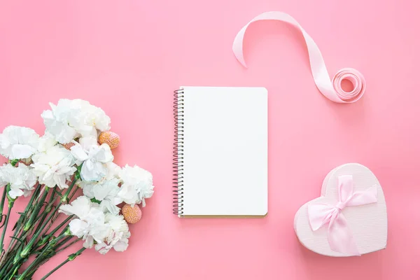 Blank notepad, bouquet of flowers and heart shaped gift box on a pink background, flat lay, copy space.
