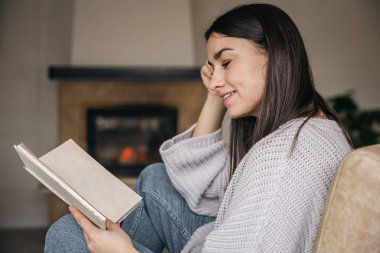Evde kitap okuyan çekici genç bir kadın. Arka planda bulanık bir şömine var..