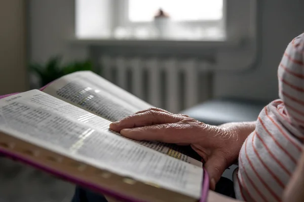 An old woman reads the Bible at home, wrinkled hands close up, copy space.