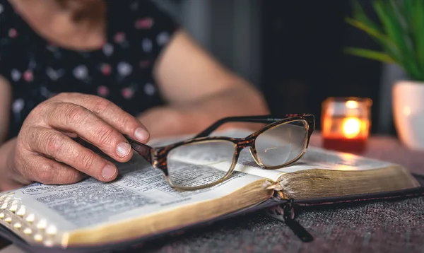 An old woman reads the Bible and holding glasses, hands close up.