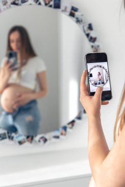 A pregnant woman takes a selfie on a smartphone in the mirror, the concept of expecting a baby.