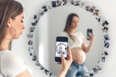 A pregnant woman takes a selfie on a smartphone in the mirror, the concept of expecting a baby.