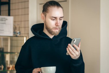 A business young man with a beard looks at the smartphone screen and drinks coffee in the morning in a cafe.