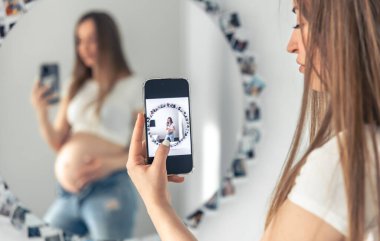 A pregnant woman takes a selfie on a smartphone in the mirror, the concept of expecting a baby.