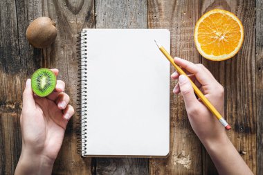 Blank notepad, female hands and and fruits on a wooden background, top view, copy space.