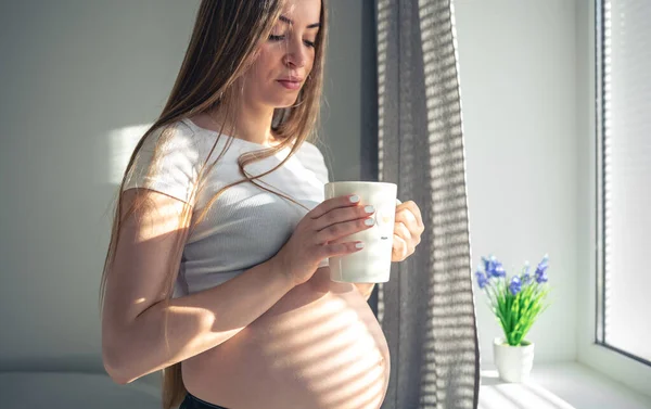 An attractive pregnant woman with a cup of tea at the window in the morning, the concept of womens health and the expectation of a child.
