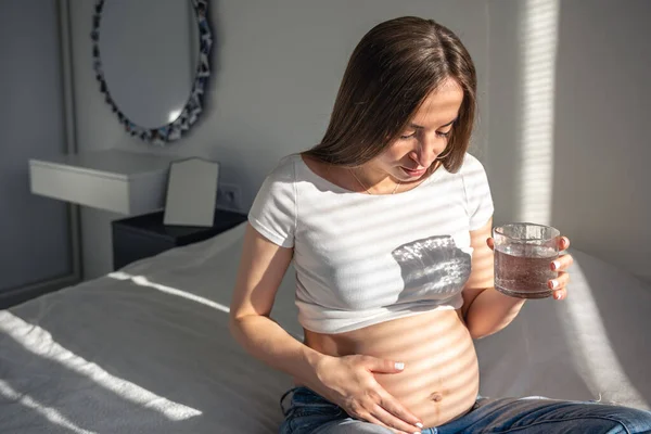 Pregnant woman with a glass of water in the room interior in the morning, health concept.