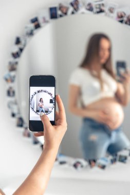 A pregnant woman takes a selfie on a smartphone in the mirror, the concept of expecting a baby.