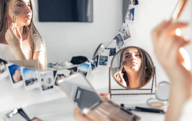 A young pregnant woman applying makeup at home in front of a mirror.