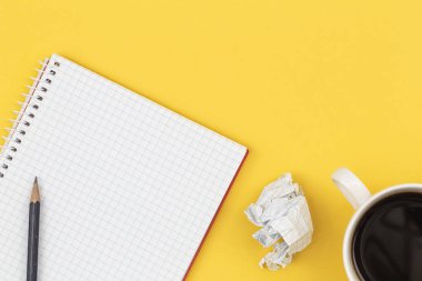 Empty notebook with cup of coffee and crumpled paper balls on a yellow background, flat lay.