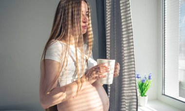An attractive pregnant woman with a cup of tea at the window in the morning, the concept of womens health and the expectation of a child.