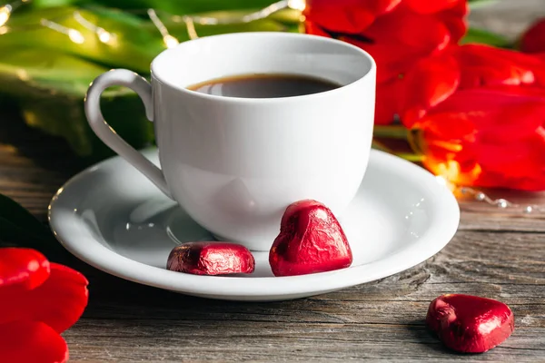 Composition with a cup of coffee, heart-shaped red foil candies and red flowers close-up.