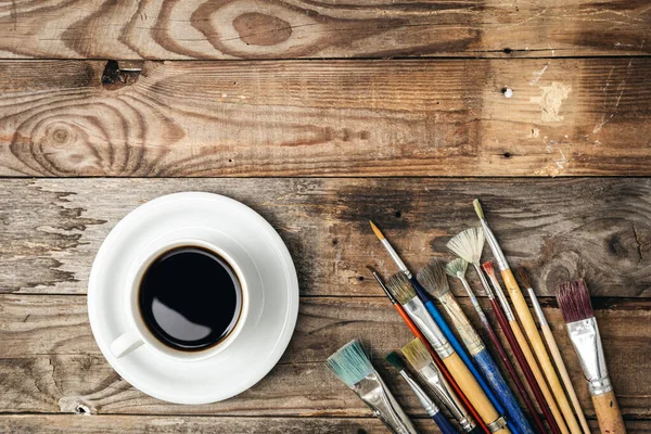 Paint brushes and a cup of coffee on a wooden background, flat lay, copy space.