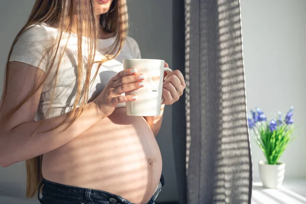 A pregnant woman with a cup of tea at the window in the morning, the concept of womens health and the expectation of a child.