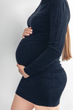 Stylish young pregnant woman in a black short dress on a white background, belly close-up.