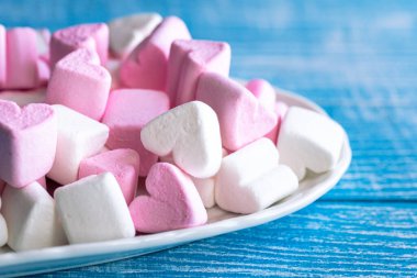 Plate with pink and white marshmallows in the form of hearts on a blue wooden background, close-up.