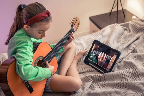 A little girl learns to play the guitar while sitting on the bed in the room, a music lesson with an online teacher, distance learning to play the guitar.