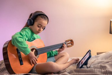 A little girl learns to play the guitar while sitting on the bed in the room, a music lesson with an online teacher, distance learning to play the guitar.