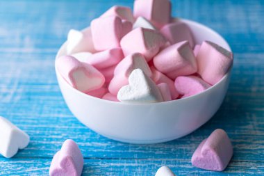 Bowl with pink and white marshmallows in the form of hearts on a blue wooden background, close-up.