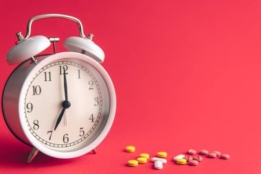 Time for health check concept. White alarm clock with pills, against the backdrop of Viva Magenta, copy space.