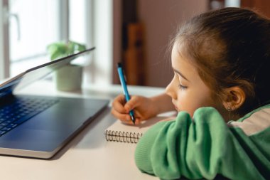 Küçük kız dizüstü bilgisayarın önünde otururken bir defter yazıyor, online öğrenim kavramı, ev okulu..