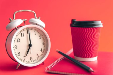 White alarm clock, paper cup and notebook on Viva Magenta blurred background, morning concept.