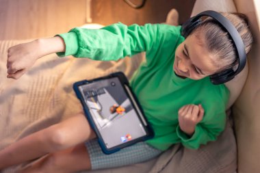 Little girl in headphones with a tablet in her room, the concept of game addiction, digitalization of children and teenagers, top view.