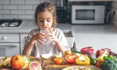 Little girl with a glass of water in a kitchen interior with fruits and vegetables.