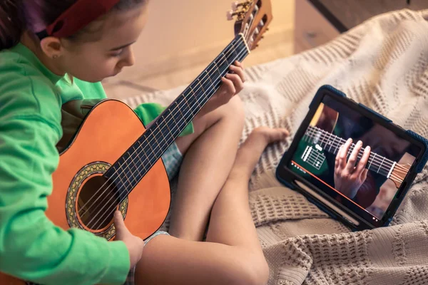 A little girl learns to play the guitar while sitting on the bed in the room, a music lesson with an online teacher, distance learning to play the guitar.