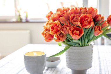 Spring composition with a bouquet of orange tulips and a candle on the table in the interior of the room.