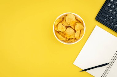 Bir tabak patates cipsi, bir hesap makinesi ve sarı arka planda bir defter, üst görünüm, beslenme ve sayma kalorisi konsepti, kopyalama alanı.