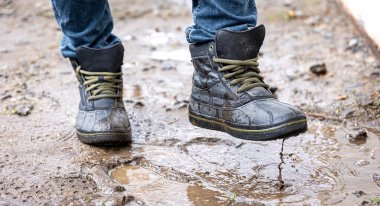 Kot pantolon ve bot giyen bir adam yağmurlu havada, kırsalda, bacakları kapalı bataklıkta yürür..