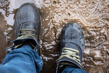 Yukarıdan bakıldığında çamurda bir çift yürüyüş ayakkabısı, kot ve bot giyen bir adam yağmurlu havada bataklıkta yürür, kapanır.