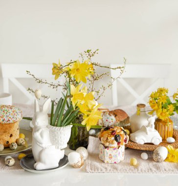 Süslü yumurtalarla Paskalya festivali masası, geleneksel pastalar ve açık arkaplanda nergis çiçekleri olan tavşan şeklinde bir vazo..