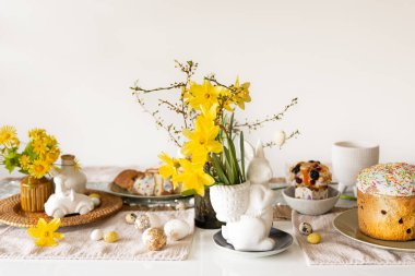 Süslü yumurtalarla Paskalya festivali masası, geleneksel pastalar ve açık arkaplanda nergis çiçekleri olan tavşan şeklinde bir vazo..