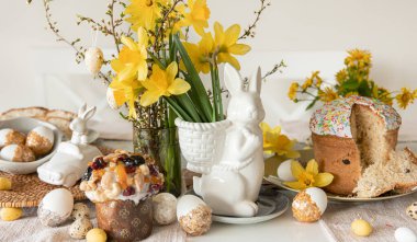 Süslü yumurtalarla Paskalya festivali masası, geleneksel pastalar ve açık arkaplanda nergis çiçekleri olan tavşan şeklinde bir vazo..