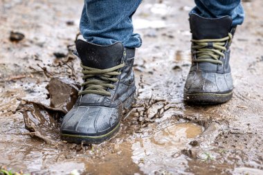 Kot pantolon ve bot giyen bir adam yağmurlu havada, kırsalda, bacakları kapalı bataklıkta yürür..