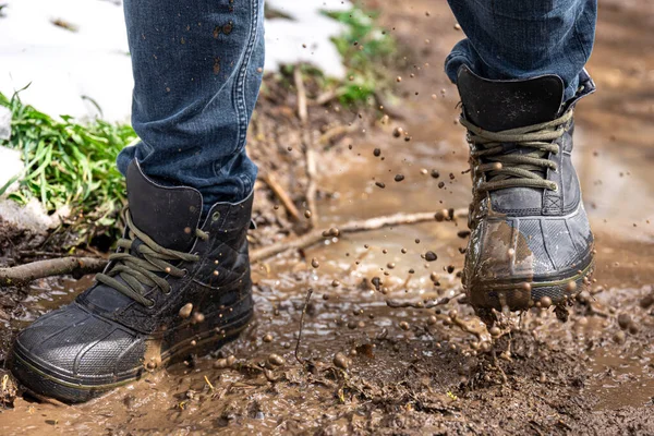 A man in jeans and boots walks through the swamp in rainy weather, in the countryside, legs close up.
