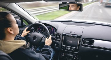 Genç, neşeli bir kadın araba kullanıyor, dikiz aynasından bakıyor..