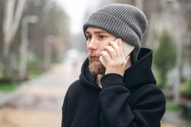 Sakallı ve şapka takan genç bir adam açık alanda bulanık bir arka planda telefonla konuşuyor..