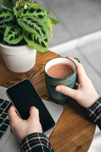 A cup of cocoa and a smartphone in the hands of a man in front of a laptop, a man works in the office, close-up.