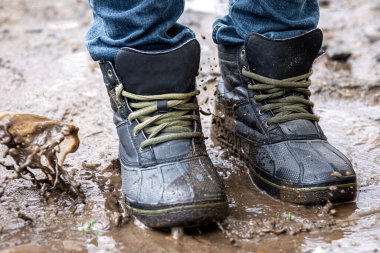 Kot pantolon ve bot giyen bir adam yağmurlu havada, kırsalda, bacakları kapalı bataklıkta yürür..