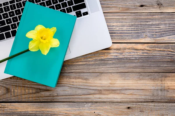 Laptop, blue book and yellow flower on wooden background, top view, workplace concept, copy space.
