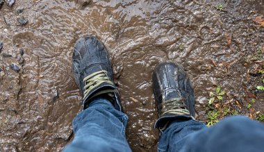 Yukarıdan bakıldığında çamurda bir çift yürüyüş ayakkabısı, kot ve bot giyen bir adam yağmurlu havada bataklıkta yürür, kapanır.